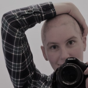 Picture of me (a white non-binary person in their 30s) touching my head and looking in the mirror while holding my camera